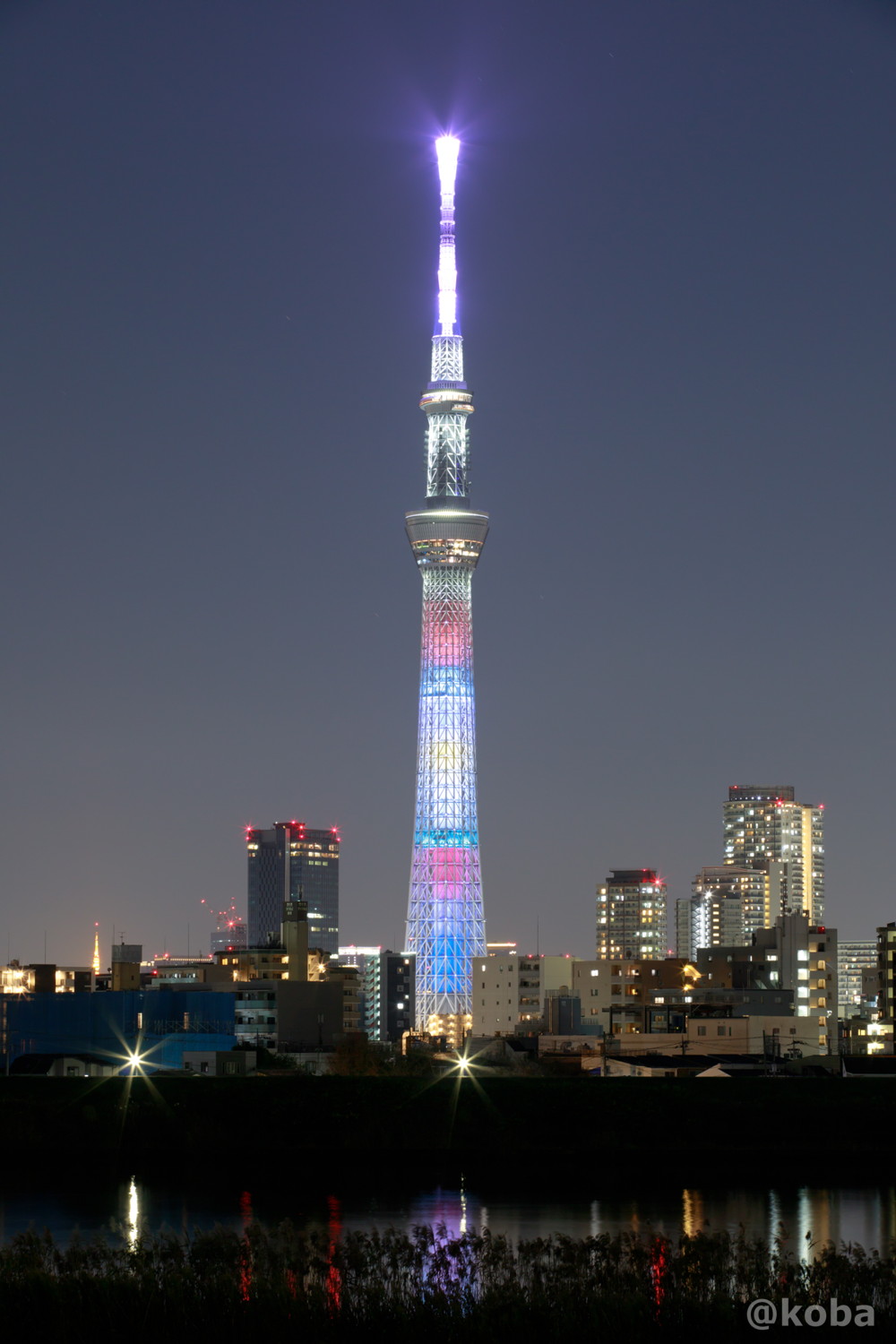 2020年デビューしたNiziU（ニジュー）の1st Album『U』のリリースを記念し、NiziUメンバーカラーの9色をあしらったライティング。 塔体に横縞に重なり合い、トップからは『U』のテーマカラーであるピンクの光が空へ放たれています。｜ライティングイメージ：NiziU　2021年11月23日｜葛飾区・四ツ木｜カメラ：Canon｜こばフォトブログ