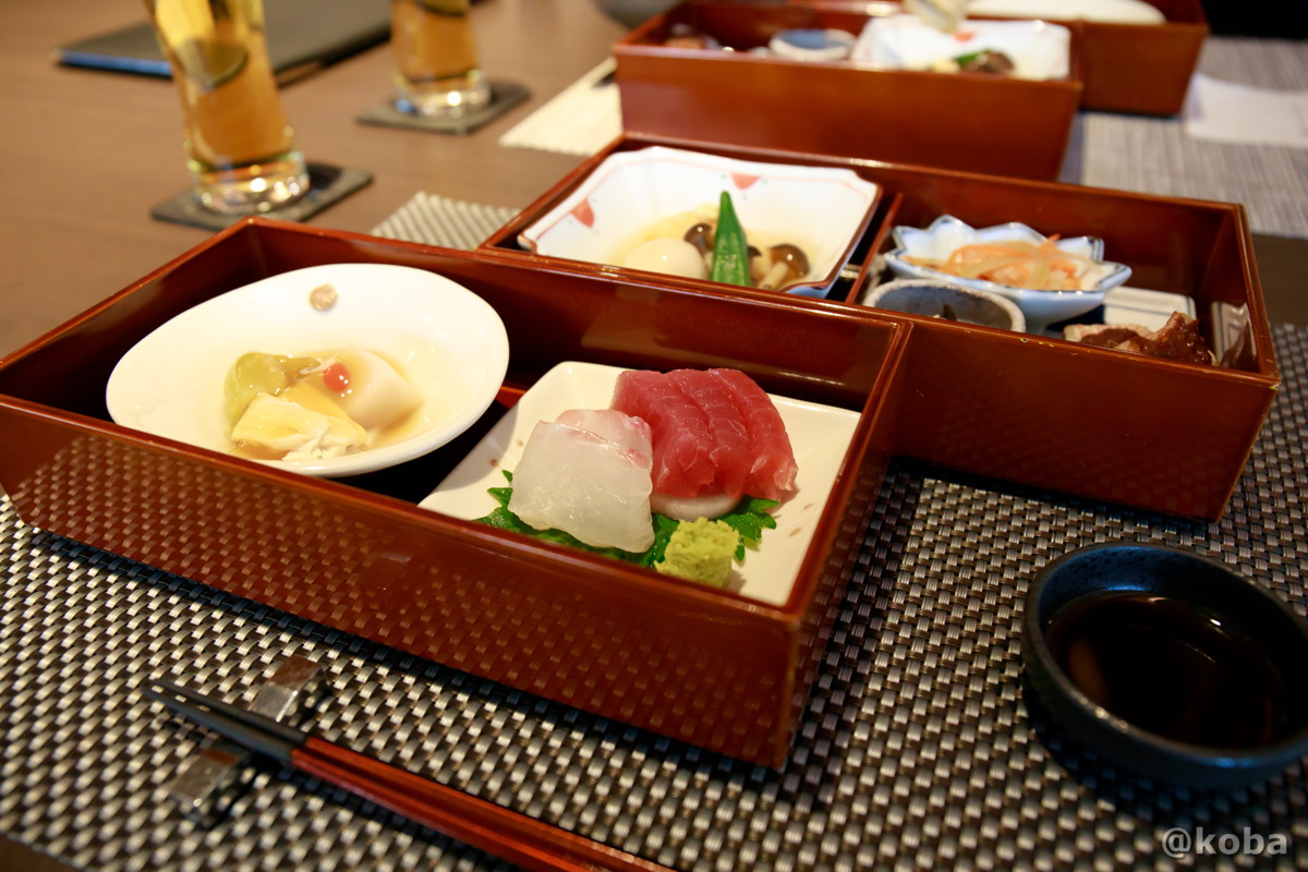 ホテルハーヴェスト勝浦の夕食。かぶら豆腐、お造り、馬鈴薯饅頭、赤魚牛蒡味噌焼などの多彩な料理が、箱の中に小分けにして美しく並べられた写真。