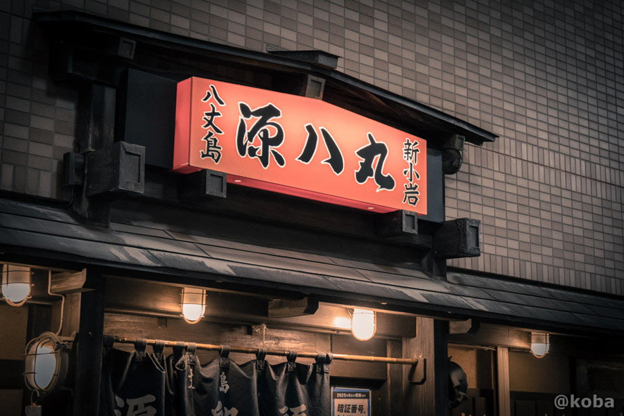 「八丈島 源八丸 新小岩店」の店名が力強く刻まれた、温かみのあるオレンジ色が特徴的なレトロな看板