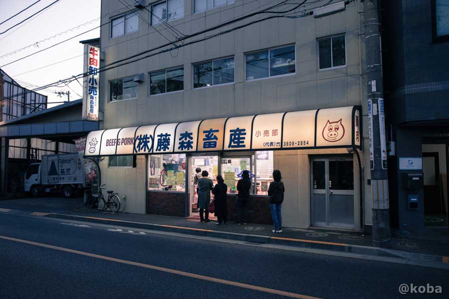 葛飾区西水元にある精肉店「藤森畜産」の外観。夕暮れ時に看板の明かりが灯り、店頭にお客さんが集まっている様子
