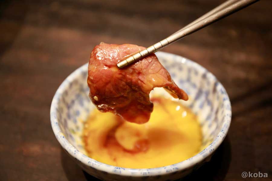 藤森畜産(葛飾区西水元)で購入した国内産牛赤身肉(ロース)を炭火で焼き、箸で濃厚な卵黄とジャンダレに浸している様子。タレと卵黄が肉に絡み、赤身肉の旨味を最大限に引き出すシズル感たっぷりのクローズアップ写真