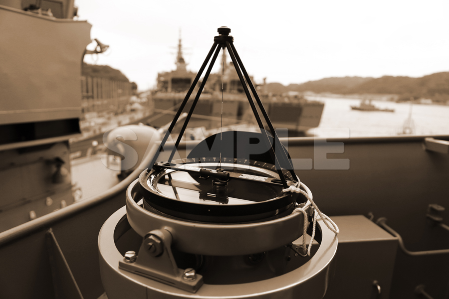 護衛艦「とね」の甲板に設置された羅針盤（コンパス）。セピア調に加工された、背景に他の艦艇や港を望むアンティークな雰囲気のミリタリー写真。

